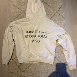 Vit hoodie Acne Studios Stockholm - Säljer en clean vit hoodie från Acne Studios med tryck på ryggen: 'Acne Studios STOCKHOLM 1996'. Tröjan har huva med snörning, långa ärmar och ribbade muddar. Perfekt för dig som gillar minimalistisk stil och svensk design.