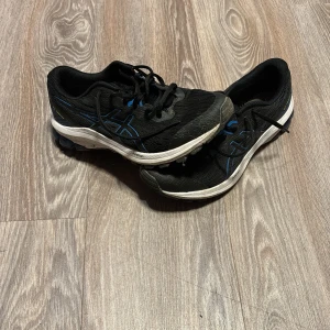 Svarta Asics sneakers med blå detaljer - Säljer ett par svarta Asics sneakers med blå och vita detaljer. Skorna har snörning, rund tå och en platt sula med gel-dämpning. Ovandelen är i syntetmaterial och mesh för bra ventilation. Perfekta för träning eller promenader.