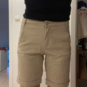 Beige midwaist shorts med uppvikta kanter - Snygga beige shorts med midwaist passform och klassisk knappstängning samt uppvikta benslut. Perfekta för varma dagar.