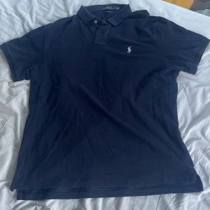 Mörkblå Polo Ralph Lauren t-shirt XL - Snygg mörkblå t-shirt från Polo Ralph Lauren i klassisk slim fit. T-shirten har korta ärmar, krage med två knappar och den ikoniska broderade loggan på bröstet. Tillverkad i 100% bomull för skön känsla och stilren look. Inga defekter. Priset kan diskuteras. 