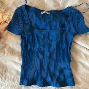 Blå ribbad topp från Zara - Snygg blå ribbad topp från Zara med fyrkantig ringning och korta ärmar. Materialet är mjukt och stretchigt, perfekt för att ge en slim passform. Toppen har en cool struktur med vertikala ribbor som ger extra stilpoäng.