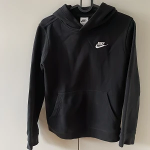 Svart hoodie från Nike med logga - Svart hoodie från Nike med klassisk vit logga på bröstet. Tröjan har huva, magficka och långa ärmar. Mjuk insida och normal passform.
