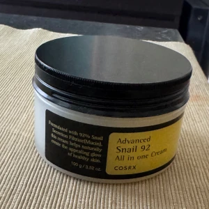 COSRX Advanced Snail 92 All in one Cream - En fuktgivande ansiktskräm från COSRX med 92% snigelsekretfiltrat. Krämen är vit och har en gel-liknande konsistens som känns lätt på huden. Kommer i en rund plastburk med svart lock och gul etikett. Perfekt för dig som vill ha glow och återfuktning.