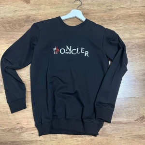 Svart sweatshirt från Moncler Paris - Svart sweatshirt från Moncler Paris med broderad logga i vitt och rött på bröstet. Tröjan har rund hals, långa ärmar och ribbade muddar vid ärmslut och nederkant. Perfekt för dig som gillar stilrena och exklusiva märken.