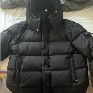 Svart Moncler pufferjacka med huva - Säljer en svart pufferjacka från Moncler med stor huva och snygga detaljer som logotyp på ärmen. Jackan har dragkedja och knappar framtill, samt praktiska fickor med dragkedja. Perfekt för kalla dagar och riktigt stilren look.