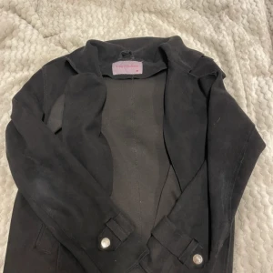 Svart mocka rock från King of Fashion - Svart rock i mocka-look från King of Fashion, storlek M. Snygg krage och coola detaljer med knappar vid ärmsluten. Jackan har en stilren och klassisk form som ger en clean vibe. Perfekt för dig som gillar minimalistisk stil och vill ha något som sticker ut.