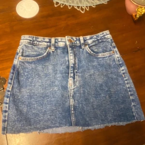 Blå jeanskjol från Bershka - Säljer en blå jeanskjol från Bershka i storlek 38. Kjolen har hög midja, klassisk femficksdesign och rå kant nedtill för en cool vibe. Materialet är tvättad denim med ljusa detaljer och slitningar. Perfekt för dig som gillar streetstyle och vill ha en snygg kortkjol.