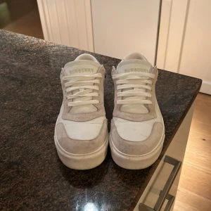 Burberry sneakers med rutig detalj - Säljer ett par snygga Burberry sneakers i vitt skinn och mocka med klassisk rutig panel i beige, svart och röd på hälen. Skorna är perfekt nu till vintern då dom är varma o mjuka. Perfekt för dig som gillar lyxiga och tidlösa detaljer.