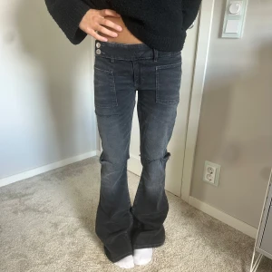 Mörkgrå bootcut jeans med fickor - Snygga mörkgrå jeans med bootcut passform och  slitningar vid knäna. Byxorna har stora fickor framtill och två silverfärgade knappar i midjan. Aldrig använda🥰🥰