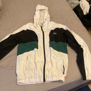 Lacoste vindjacka med luva  - Snygg vindjacka från Lacoste i vitt, svart och grönt. Jackan har huva, dragkedja framtill och två fickor med dragkedja. Klassisk Lacoste-logga på bröstet och ärmen. Perfekt för dig som gillar sportig och clean stil.