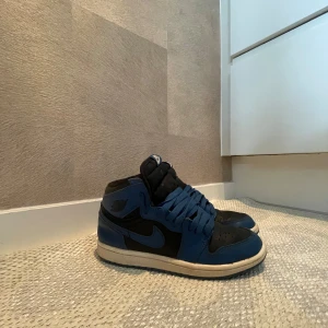 Nike Jordan 1 Mid blå/svarta sneakers storlek 33 - Nike Jordan 1 Mid sneakers i blått och svart med klassisk snörning och rund tå. Skorna har en vit mellansula och blå yttersula, samt ikoniska Jordan Wings-loggan på sidan. Tillverkade i skinn och syntetmaterial för en schysst look. Perfekta för dig som gillar streetwear och sneakers. I bra andvent skick 👌👍💯