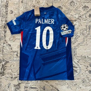 Chelsea Palmer #10 Nike matchtröja - Chelsea FC matchtröja med Palmer och nummer 10 på ryggen. Snyggt geometriskt mönster i marinblått och blått, med vita och röda detaljer vid sidorna. UEFA Foundation-märke på ärmen och klassisk Nike-logga framtill. Tillverkad i lätt, ventilerande polyester.