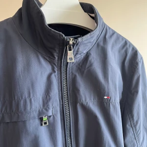 Tommy Hilfiger Vindkacka  - Snygg mörkblå vindjacka från Tommy Hilfiger i storlek M. Jackan har dragkedja framtill, ståkrage och flera fickor med dragkedja. Liten broderad logga på bröstet och diskret design som passar till det mesta. Perfekt för dig som gillar stilrena plagg med sportig känsla.