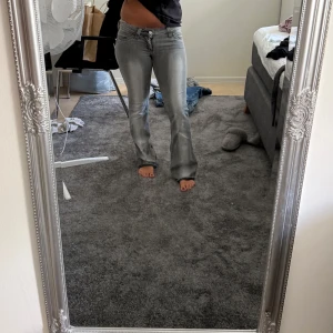 Grå lågmidjade bootcut jeans med låg midja - Säljer ett par grå jeans med bootcut och låg midja. Jeansen har klassisk femficksdesign, dragkedja och knapp framtill samt en cool grön patch bak i midjan. Snygg tvätt och stretchigt material som sitter tajt upptill och släpper ut vid benen.