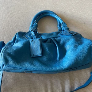 Blå handväska från Marc by Marc Jacobs - Snygg blå handväska från Marc by Marc Jacobs i mjukt skinn med coola detaljer och dubbla handtag. Väskan har en avtagbar axelrem och dragkedja upptill. Insidan är fodrad med svartvitt tryck och har innerfack. Perfekt storlek för allt du behöver.