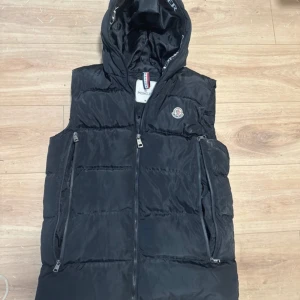 Svart dunväst från Moncler med huva - Svart dunväst från Moncler med avtagbar huva och klassisk logga på bröstet. Västen har flera dragkedjeförsedda fickor och quiltad design. Perfekt för lager-på-lager och streetstyle. Insidan har ett coolt serietidningsmönster med tvättråd.