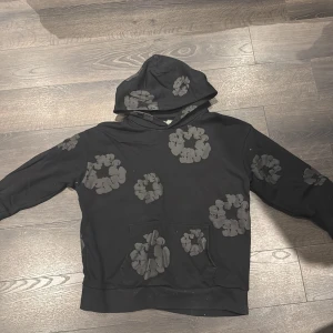 Svart hoodie med blommor från Denim Tears - Svart hoodie från Denim Tears med coola blommiga mönster i mörkgrått över hela tröjan, inklusive huvan och ärmarna. Tröjan har en klassisk känguruficka framtill och en relaxed passform. Perfekt för dig som vill sticka ut med en unik streetwear-look.