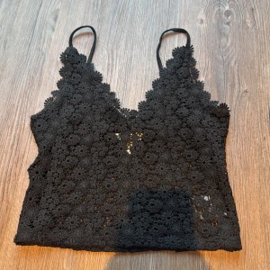 Svart spetsig linnetopp från H&M - Svart linnetopp från H&M med tunna axelband och blommig spets över hela toppen. V-ringad och croppad modell med snygga spetsdetaljer längs kanterna. Perfekt att styla med höga jeans eller kjol för en cool look.