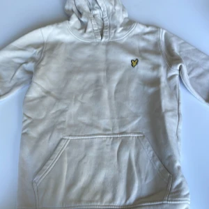 Vit hoodie från Lyle & Scott Junior - Snygg vit hoodie från Lyle & Scott Junior med klassisk gul logga på bröstet. Tröjan har huva, känguruficka och långa ärmar. Mjuk bomullskvalitet som är skön att ha på sig. Perfekt för chill dagar eller när du vill ha en clean look. Pris går att diskuteras 