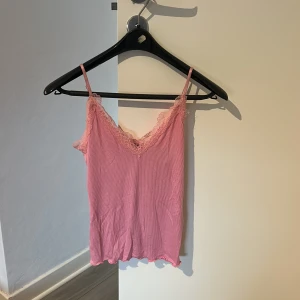 Rosa ribbad linnetopp med spets - Säljer en ljusrosa linnetopp med tunna axelband och v-ringad hals. Toppen har ribbad struktur och är dekorerad med romantisk spets längs halsringningen och ryggen. Perfekt för dig som gillar söta detaljer och en feminin vibe.