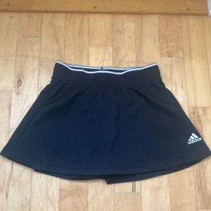 Svart sportkjol från Adidas - Svart kortkjol från Adidas med inbyggda shorts och elastisk midja med vit rand. Materialet är lätt och andas, perfekt för tennis eller träning. Diskret Adidas-logga i vitt framtill och Aeroready-tryck bak. Snygg och funktionell för aktiva dagar.