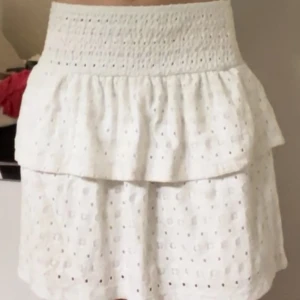 Vit volangkjol - Supersöt vit kortkjol med bred smock i midjan och dubbla volanger. Kjolen har broderade detaljer och små hålmönster. Perfekt för sommardagar