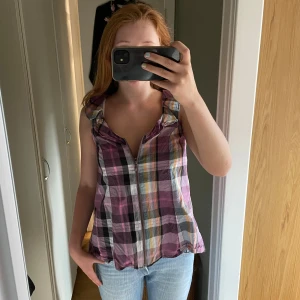 Rutig ärmlös blus med dragkedja - Säljer en luftig och färgglad rutig blus i lila, rosa, vit och gul. Toppen är ärmlös med en liten volang vid axlarna och har dragkedja framtill. Perfekt för varma dagar och enkel att styla med jeans eller shorts.