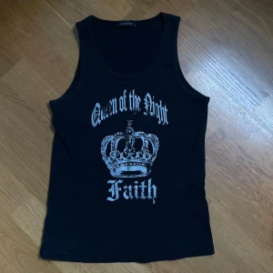Svart ribbad linnetopp med silver tryck och strass stenar  - Svart ribbad linnetopp från b.young med silverfärgat tryck som visar en krona och texten 'Queen of the Night Faith' i gotisk stil. Modellen är ärmlös och har rundad hals. Perfekt för dig som gillar edgy och statement-plagg.