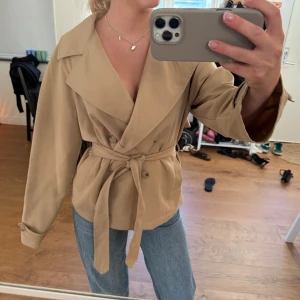Beige trenchcoat från Stradivarius XS - Snygg beige trenchcoat från Stradivarius i storlek XS. Jackan har bred krage, dubbelknäppning och ett bälte i midjan som ger en schysst siluett. Långa ärmar med knappdetalj vid mudden