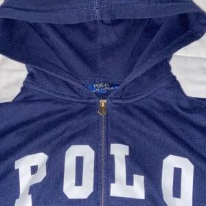 Mörkblå hoodie från Polo Ralph Lauren - Säljer en mörkblå hoodie från Polo Ralph Lauren med stor vit POLO-text på bröstet. Tröjan har huva, dragkedja framtill och är tillverkad i mjuk bomull. Perfekt för chill dagar och har en klassisk streetwear-vibe.