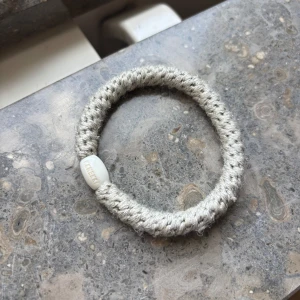 Beige stickat armband - Snyggt beige stickat armband med en oval vit pärldetalj där märket är ingraverat. Armbandet har en grov, stickad struktur som ger en cool och unik look. Perfekt för dig som gillar enkla men stilrena accessoarer.