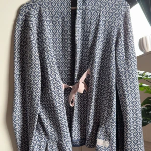 Blåvit cardigan jacka Odd Molly - Snygg blåvit cardigan jacka från Odd Molly med långärmad design och ett mönster av hjärtan. Jackan har en rosa knyt framtill och volangdetalj baktill. Mjuk stickad känsla, perfekt för dig som gillar unika detaljer och skön stil.