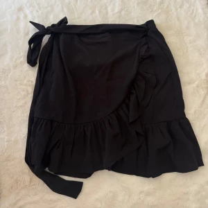 Svart omlottkjol med volang från ONLY - Svart kort omlottkjol från ONLY i storlek L. Kjolen har volangkant och knytband i midjan som ger en snygg och feminin look. Perfekt för dig som gillar stilrena plagg med lite extra detalj. Materialet känns lätt och skönt mot huden.