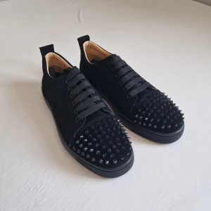 louboutins - Säljer ett par svarta sneakers från Louboutin med coola svarta nitar på tån och klassisk röd sula. Skorna har snörning och är tillverkade i mocka med en platt sula. Insidan är beige och de har en rund tå. Perfekt för dig som vill sticka ut med din stil. Storlek 43 2/3