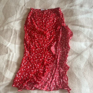 Röd blommig omlottkjol från Bershka - Säljer en röd långkjol från Bershka med blommigt mönster och omlott-design. Kjolen har volangdetaljer längs kanten och är gjord i ett lätt och luftigt material som passar perfekt till varma dagar. Supersnygg att matcha med en enkel topp och sneakers.