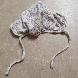 Mössa - Söt mössa från Newbie i mjuk bomull med blommigt mönster i rosa och grönt på vit botten. Mössan har praktiska knytband under hakan och en lätt rynkad form som ger en gullig look. Perfekt för dig som gillar romantiska detaljer och mjuka material.