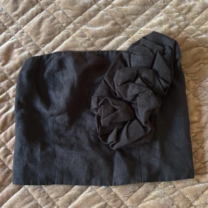 Svart bandeau tubtopp med rosett - Säljer en svart bandeau tubtopp med en stor, cool rosett på ena sidan. Toppen har smockad baksida och dragkedja för enkel på- och avtagning. Materialet känns som linne och toppen är axelbandslös, perfekt för att skapa en edgy look.