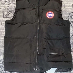 Svart dunväst från Canada Goose - Säljer en svart dunväst från Canada Goose med knappar och dragkedja. Västen har två stora fickor framtill och en broderad logotyp på bröstet. Perfekt för kyliga dagar. Pris kan diskuteras vid snabb affär
