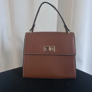 Brun handväska med metallspänne - Snygg brun handväska i skinn med ett elegant metallspänne framtill. Väskan har en enkel och stilren design med en kort handrem. Perfekt för att ge en klassisk touch till din outfit. Knappt använd