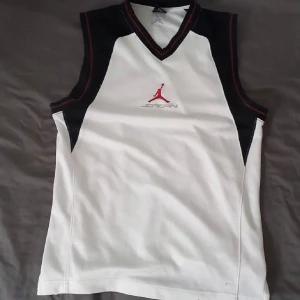 Vit ärmlös Jordan-tröja - Säljer en vit ärmlös tröja från Jordan med svarta och röda detaljer. Tröjan har en v-ringad hals och är perfekt för sportiga aktiviteter. Den är tillverkad i Thailand och har en stilren design med det ikoniska Jordan-logotypen på framsidan.