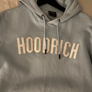Ljusblå hoodie från Hoodrich - Säljer en snygg ljusblå hoodie från Hoodrich med stor vit logga på bröstet. Hoodien har en justerbar huva med dragsko och ribbade muddar vid ärmslut och nederkant. Perfekt för en avslappnad stil.