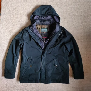 Barbour field jacka - Säljer en Barbour windbreaker/field jacka i nyskick. Väldigt stilren design med luva som sticker ut från de flesta andra Barbour jackor. Jackan är en längre version. Hål i ena fickan. Skriv vid minsta fundering! 