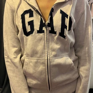 Grå hoodie från GAP - Säljer en klassisk grå hoodie från GAP med dragkedja och stor logga på framsidan. Tröjan har en bekväm passform med långa ärmar och en praktisk huva. Perfekt för en avslappnad stil.