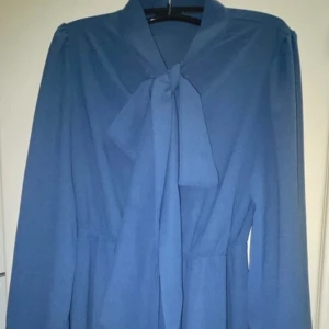 Blå klänning med knyt - Elegant blå klänning med långa ärmar och knyt i halsen. Perfekt för en stilren look.