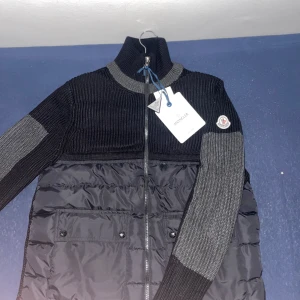 Svart och grå jacka från Moncler - Snygg svart och grå jacka från Moncler med stickade ärmar och quiltad kropp. Jackan har en dragkedja framtill och en hög krage. Perfekt för kyligare dagar med sin stilrena design och praktiska fickor med knappar.