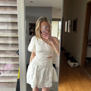 Assymetrisk studentklänning - Supersöt studentklänning/vit mini eller sommarklänning. Enärmad puffärm och en liten cutout på sidan, lite blomm-aktigt mönster. Använd ett par gånger men super skick! Nypris 400kr. True to size. Bara stt skriva vid frågor🥰