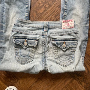 Ljusblå jeans med slitna detaljer - Lågmidjade bootcut jeans från True Religion i en så fin ljusblå tvätt. Jag är 169 cm lång. Jeansens defekter är de som syns på bilderna. Bild 5 är på insidan av jeansen, alltså inget som syns utifrån, och det är jag som har lagat/förebyggt skador. Dessa har varit mina favorit jeans väldigt länge och jag säljer dom bara för bra bud!❤️
