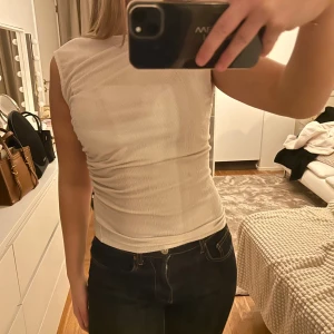 Beige ribbad topp - Säljer en stilren beige ribbad topp med tight passform. Toppen är ärmlös och har en snygg rynkdetalj på sidan. Perfekt för en trendig look!