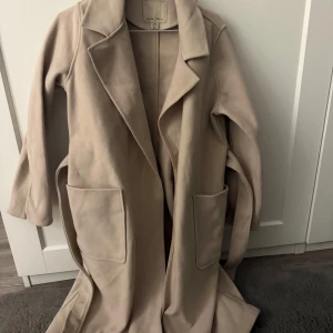 Beige kappa från Amisu - Säljer en stilren beige kappa från Amisu i storlek EU 38. Den har långa ärmar och ett bälte i midjan för en snygg passform. Perfekt för höst och vår med sina stora fickor och klassiska design.