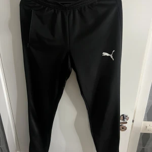 Svarta träningsbyxor från Puma - Snygga svarta träningsbyxor från Puma med elastisk midja och dragkedja vid fickorna. Perfekta för träning eller en avslappnad dag. Puma-loggan syns tydligt på benet.